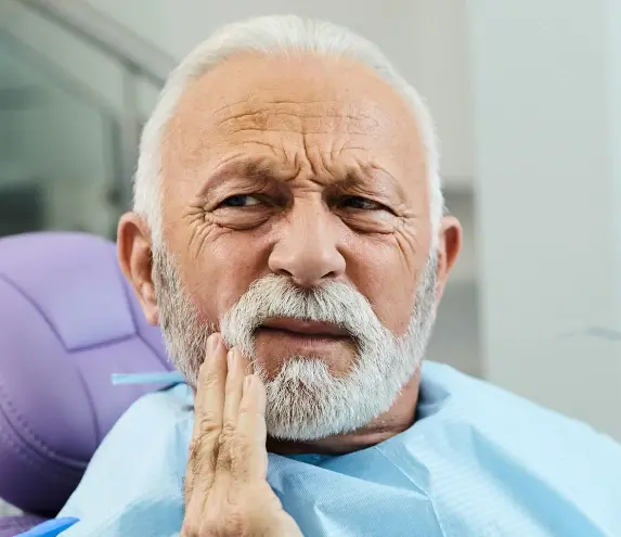Older man experiencing tooth pain, holding his cheek in a dental office setting.