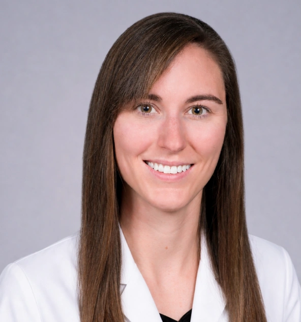Professional headshot of female dentist smiling, wearing white coat against gray background.